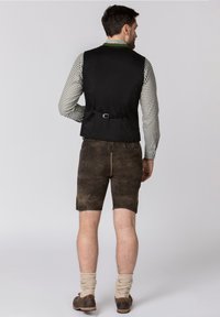 Gilet nero con una chiusura regolabile sul retro, abbinato a pantaloni corti marroni. I pantaloni corti hanno una finitura testurizzata, arrivano fino al ginocchio e sono abbinati a calze lavorate di colore beige.