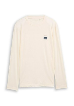 Pikkade varrukatega t-särk helevalgest kangast, millel on sile tekstuur. Rinnal on väike, tumesinise värvi ristkülikukujuline silt.