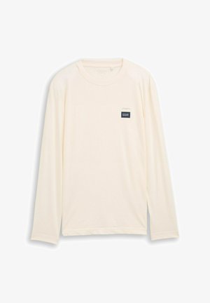 Pikkade varrukatega t-särk helevalgest kangast, millel on sile tekstuur. Rinnal on väike, tumesinise värvi ristkülikukujuline silt.