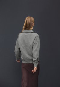 Pull-over gris avec col et coupe décontractée, associé à une jupe marron foncé. Texture douce et design minimaliste.