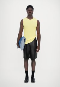 Kollane sooneta top, mustad nahast lühikesed püksid, sinine teksajakk, mis on käe peal, mustad sokid ja mustad kingad. Lihtne valge taust.