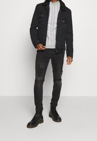 Homme portant une veste à col sherpa noire, un pull rayé gris, un jean skinny noir déchiré et des bottes noires à lacets, debout devant un fond blanc.