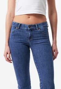 Vaqueros de denim azul ajustados con una cintura de media altura, cierre de botón y diseño de cinco bolsillos. Presenta un ligero desgastado y costuras en tono.