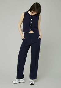 Débardeur navy sans manches avec des boutons dorés, associé à un pantalon large navy et des baskets blanches. Tissu lisse avec un design structuré.