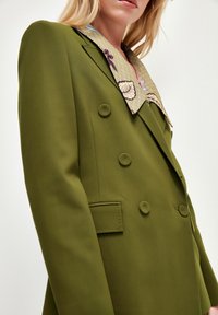 Dubbelknäppt olivgrönt blazer i en texturerad tyg, med en mönstrad krage i beige, blå och lila accenter.