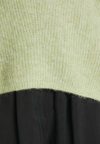 Hellgrüner gestrickter Pullover mit einer weichen, flauschigen Textur, kombiniert mit einem dunklen schwarzen Stoff darunter, der einen geschichteten Look zeigt.