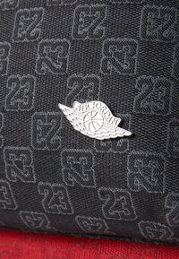 Černá texturovaná tkanina s vzorovaným designem čísla "23" v různých umístěních, doplněná stříbrnou pinovou značkou "Air Jordan".