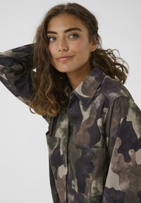 Giovane donna con capelli ricci che indossa una camicia a maniche lunghe con motivo mimetico, in posa con un braccio sollevato e un sorriso leggero.