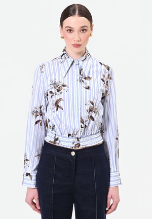 Femme aux cheveux foncés portant un chemisier à manches longues rayé et fleuri avec une taille smockée et un pantalon taille haute sombre, face à l’avant sur fond blanc.