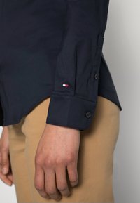 Navy long-sleeve shirt featuring buttoned cuff and a small red, white, and blue embroidered logo on the sleeve. Smooth fabric detail.