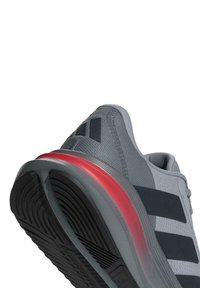Scarpa da corsa grigia con accenti neri, tomaia testurizzata e suola con tintura rossa. Presenta un motivo di battistrada distintivo e dettagli di marca.