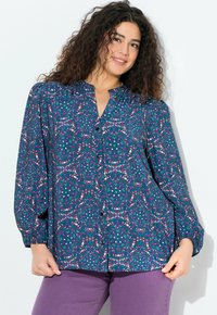 Langsleeve blouse in blauw met een kleurrijk abstract patroon. Voorzien van een knoopsluiting aan de voorkant en een geplooide halslijn. Gecombineerd met paarse broek.
