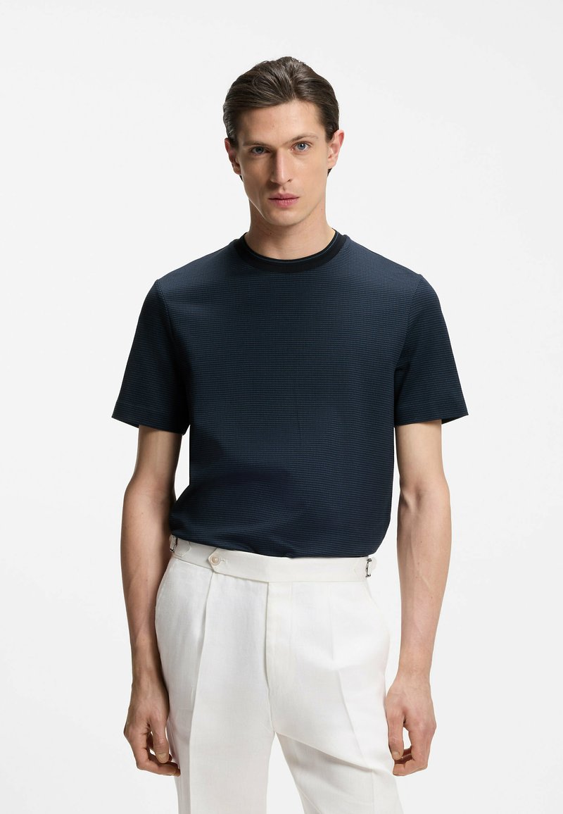 Homme aux cheveux plaqués en arrière portant une chemise texturée à manches courtes foncée et un pantalon blanc taille haute sur un fond uni.