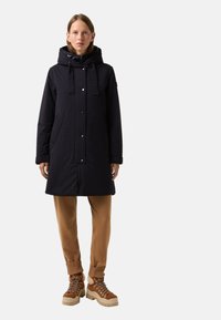 Czarna ocieplana parka z kapturem, na zatrzaski, z bocznymi kieszeniami; połączona z beżowymi, prążkowanymi spodniami i brązowymi, wiązanymi botkami z ozdobnym sprzętem.