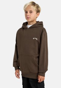 Barn med blondt hår iført en brun hettegenser med en logo, stående mot en enkel hvit bakgrunn, med hendene langs siden.