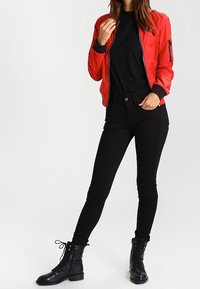 Chaqueta bomber roja con puños acanalados, camiseta negra, jeans ajustados negros y botas de combate negras con cordones. Tejido suave, diseño elegante.