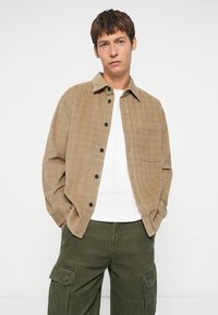Camisa abotonada marrón texturizada con un solo bolsillo, llevada sobre una camiseta blanca, combinada con pantalones de cargo de pana verde.