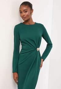 Žalia suknelė ilgomis rankovėmis, su sukimo detalėmis liemenyje ir auksine akcentacija. Lygus audinys su prigludusia siluetu. Paprastas apvalus iškirpimas.