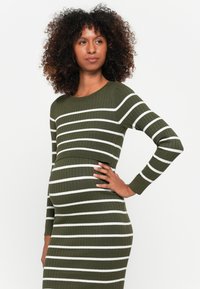 Donna incinta con capelli ricci che indossa un abito a coste verde oliva con strisce bianche orizzontali, in posa con una mano sulla vita.