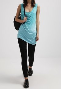 Una canotta turchese drappeggiata realizzata in tessuto liscio, abbinata a leggings neri e scarpe nere con lacci, tenuta con una borsa nera.