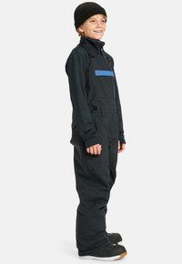 Sorte sn overaller med blå accentstribe, lavet af vandafvisende materiale. Har justerbare stropper og lynlåslommer. Sort baselag og hat.