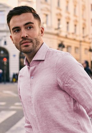 Jonge man met kort donker haar en baard, gekleed in een lichtroze overhemd met knopen, staand buiten in een stedelijke omgeving.