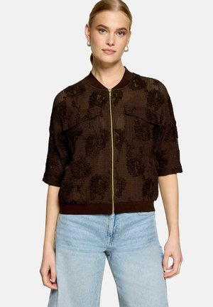 Femme portant une veste zippée marron foncé texturée à manches mi-longues et un jean bleu clair, debout devant un fond uni clair.
