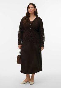 Cardigan en dentelle marron foncé avec fermeture à boutons et manches longues, associé à une jupe plissée marron foncé et des chaussures beige à enfiler.