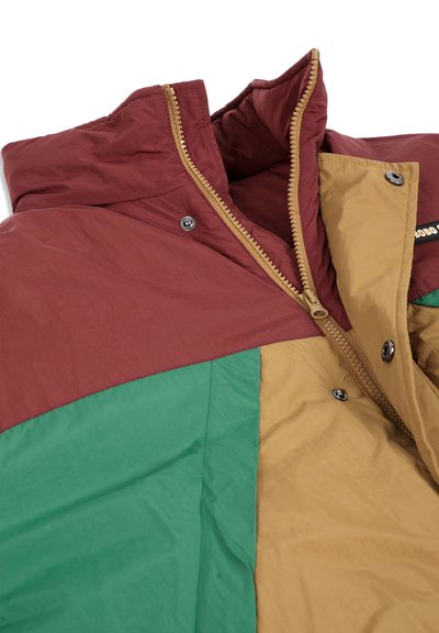 Gros plan d'une veste multicolore avec un motif en blocs de couleurs vert, marron et beige, avec une fermeture éclair et des boutons-pression.