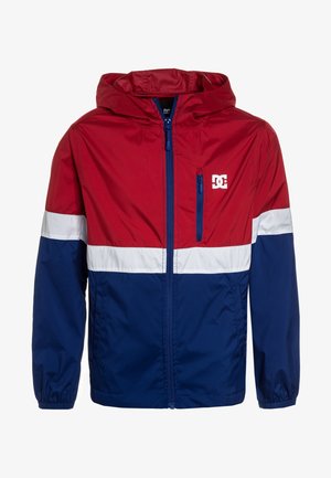 Giacca a vento rossa e blu navy con cappuccio, caratterizzata da strisce orizzontali bianche e una tasca sul petto con zip. Realizzata in materiale leggero.