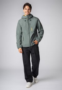 Lichtgroene, gevoerde jas met capuchon, een ritssluiting aan de voorkant, twee zijzakken en Urban Edition branding. G gedragen met zwarte jeans en witte sneakers.