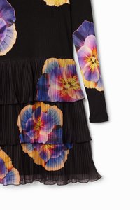 Robe noire ornée de motifs floraux multicolores vibrants, avec des textures plissées et des couches à volants. Manches longues et corsage ajusté.