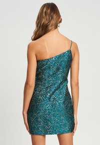 Tyrkis enaxlad klänning med en åtsittande silhuett, som har ett metalliskt mönster och en slät yta. Längd över knäna och smala band.