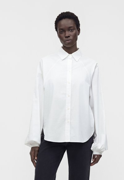 Femme aux cheveux courts portant une chemise blanche ample boutonnée avec de longues manches bouffantes et un pantalon noir, debout devant un fond uni.
