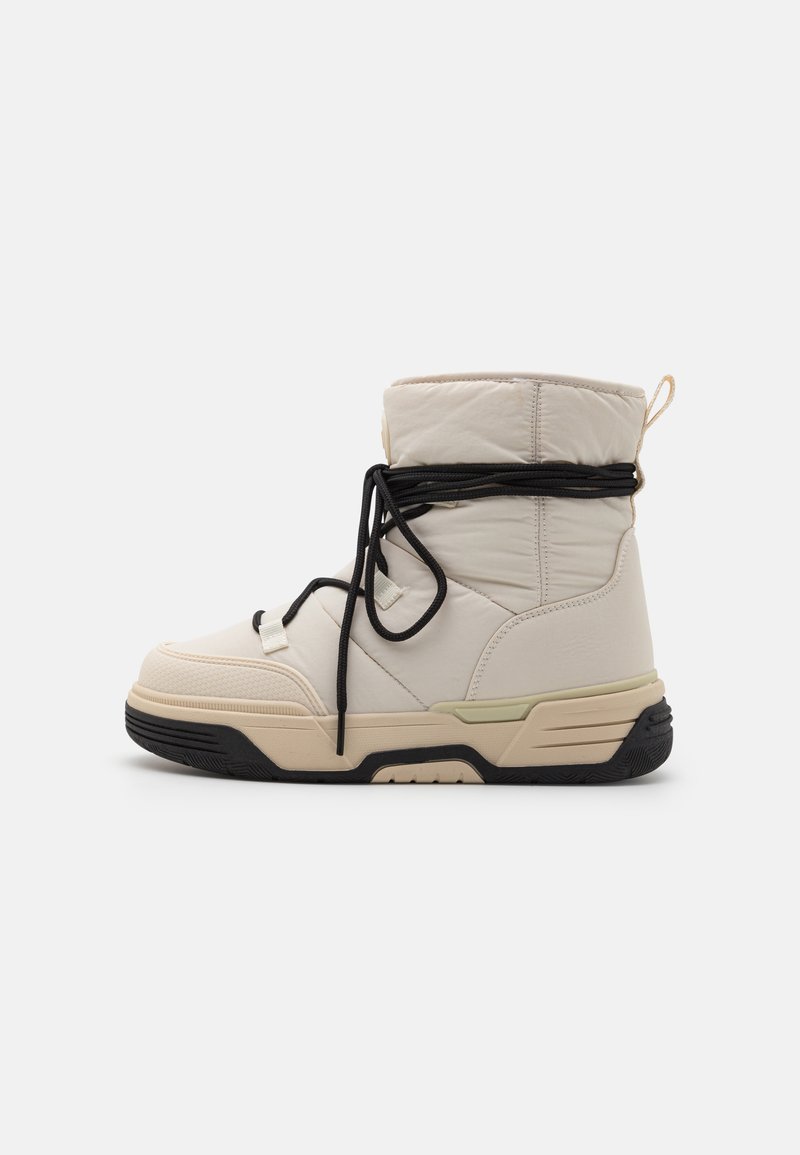 Beige winterboot met hoge gewatteerde schacht, zwarte veters om de enkel gewikkeld, dikke beige en zwarte zool, en een handgreep aan de hiel.