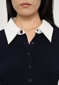 Gros plan sur une personne portant un haut côtelé bleu marine avec un grand col pointu blanc et des boutons-pression noirs à l'avant.