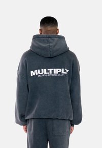 Felpa grigia oversize con cappuccio regolabile, caratterizzata da un grande logo bianco "MULTIPLY" sul retro e un tessuto morbido e testurizzato.