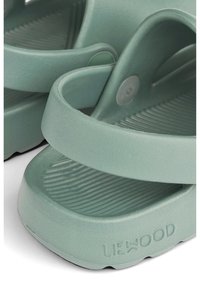 Vihreät kumiset sandaalit, joissa on kuvioitu pohja, kaksi leveää remmiä ja pyöreä koriste. Sivulla näkyy merkin nimi "LIEWOOD".