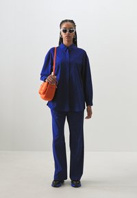 Plava dugmadna košulja dugih rukava i odgovarajuće hlače; upareno s narančastom torbom preko ramena i crnim tenisicama s plavim detaljima.