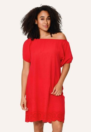 Vrouw met krullend haar die een rode off-shoulder jurk met een zoom van opengewerkte kant draagt, staand tegen een effen lichte achtergrond.