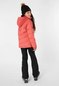 Giacca puffer corallo con cappuccio e dettagli a coste, abbinata a pantaloni da neve neri e un berretto in maglia nero con pom-pom in pelliccia sintetica.