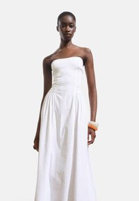 Witte strapless jurk met geplooide buste, vloeiende rok en gestructureerde stof. Inclusief oranje en witte armband om een pols.