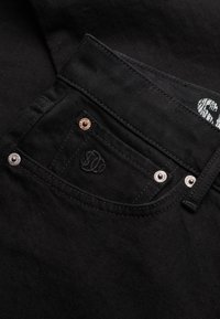 Close-up van een zwarte denim spijkerbroekzak met metalen klinknagels en geborduurd logo, met gedetailleerd stiksel en stofstructuur.