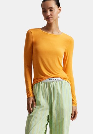 Femme portant un haut orange à manches longues et un pantalon ample à rayures vert clair avec une ceinture Ralph Lauren visible, debout devant un fond clair.