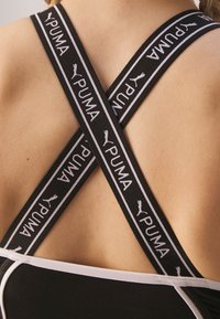 Svart atletisk linne med korsade elastiska band och vit "PUMA"-logotyp, samt en kontrasterande vit kant längs nacklinjen.