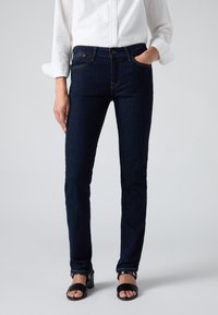 Femme portant un jean slim bleu foncé, une chemise blanche à manches longues avec poignets retroussés et des sandales noires à talons ouverts.