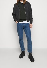 Chaqueta bomber negra sobre una sudadera con capucha verde oscuro, jeans azules y botas negras con cordones, todo sobre un fondo blanco liso.