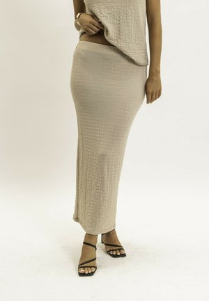 Femme portant une longue jupe texturée beige et un haut assorti, avec des sandales à talons noires à lanières, sur un fond clair uni.