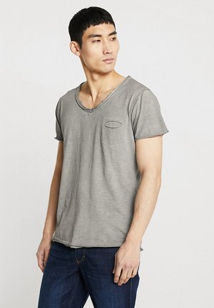 Giovane uomo con maglietta grigia a maniche corte e scollo a V e jeans blu scuro che guarda a sinistra su sfondo bianco semplice.