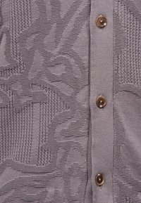 Κοντινή λήψη γκρι πλεκτού υφάσματος με υφασμένα αφηρημένα μοτίβα και τρία καφέ κουμπιά τοποθετημένα κάθετα.